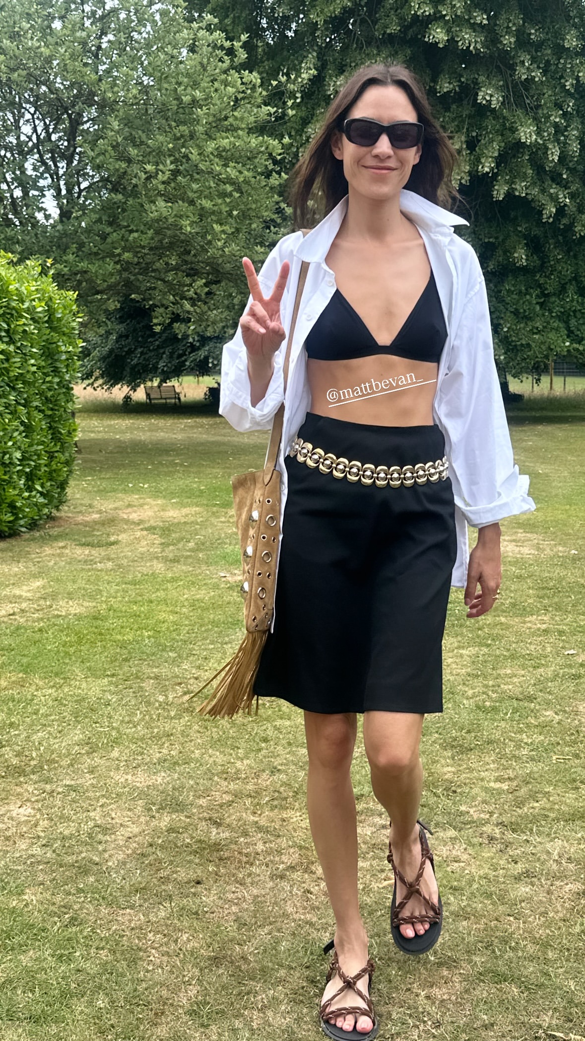 @alexachung walks along a grassy field wearing a black knee-length skirt with a black bra, white shirt, gold chain belt and brown rope sandals.