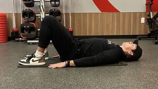 Man lies on his back on exercise mat with knees bent and feet flat on the floor. He wears Nike trainers, black pants, black sweatshirt and black cap.