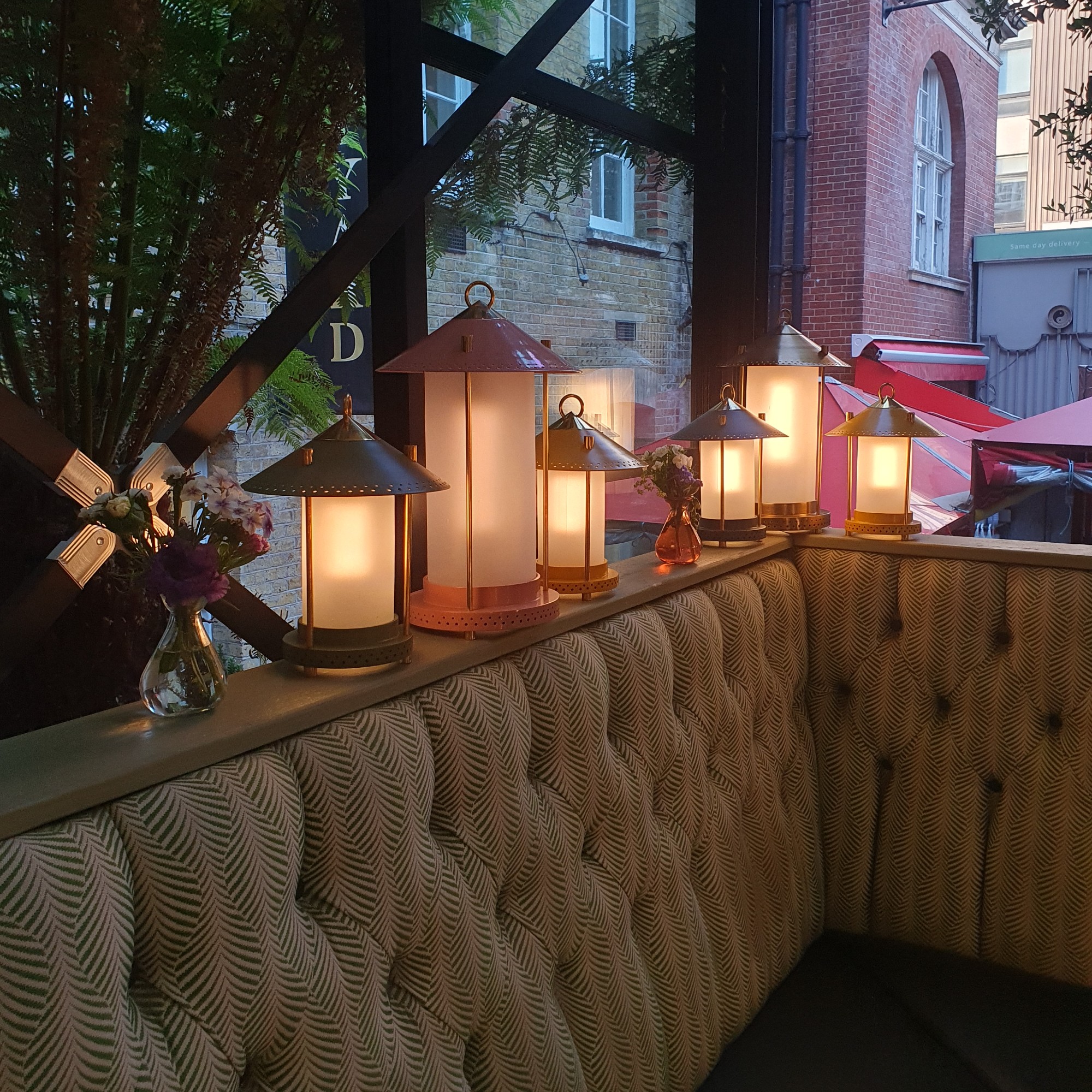 The Pooky Stefan lanterns by The Novogratz in different sizes lit up and lined up by a window on top of banquet seating