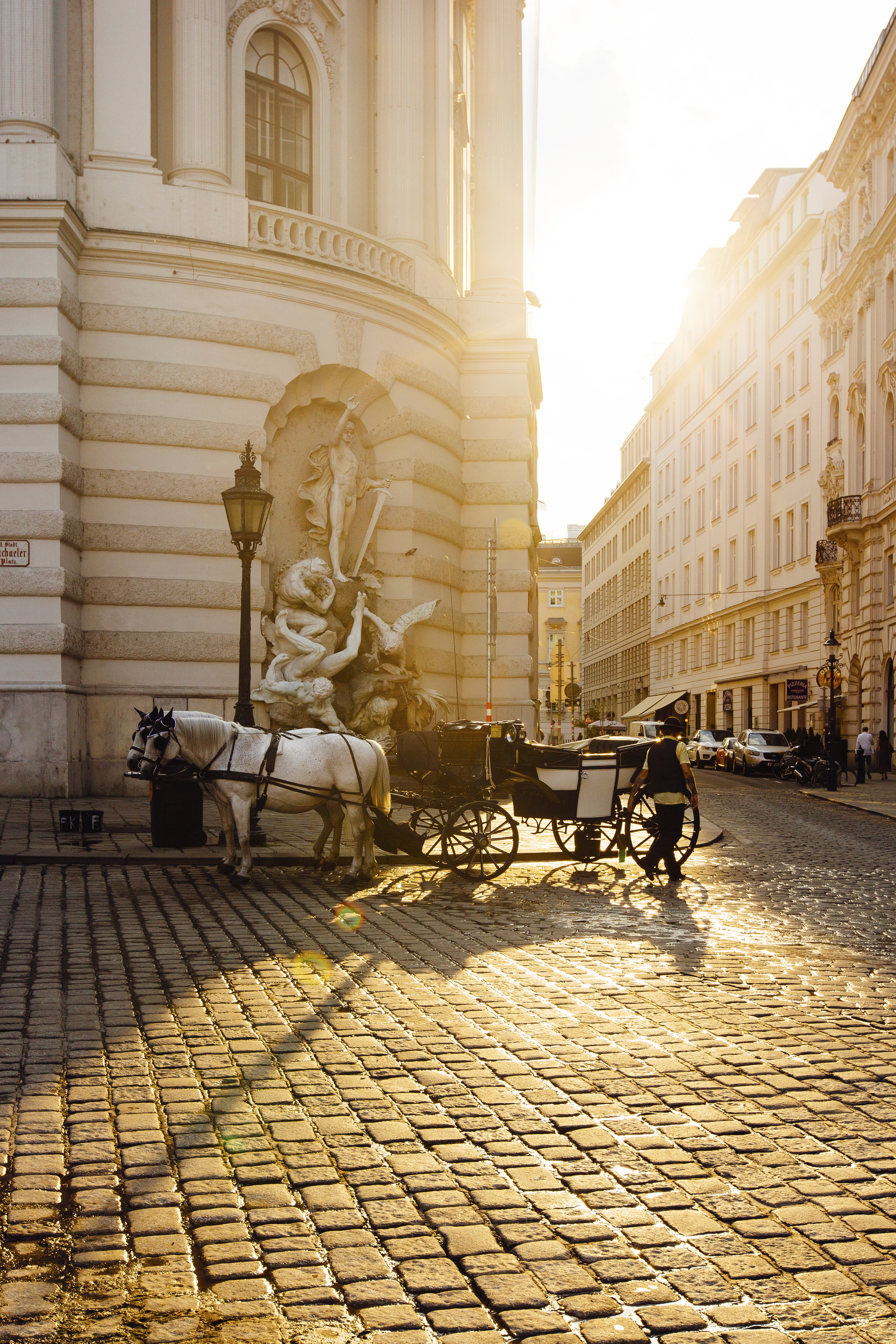 A sunlit view of a city with cobblestoned streets, horse-led carrages, and historical architecture.