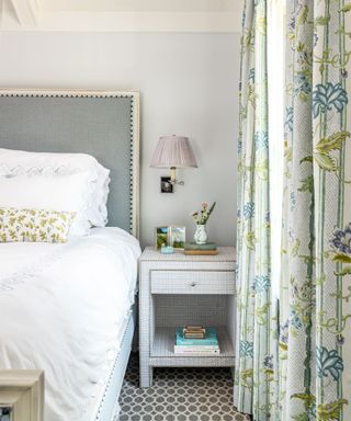 corner of bedroom with white bedding, gray headboard, neutral wall with white nightstand and blue and green patterned curtains