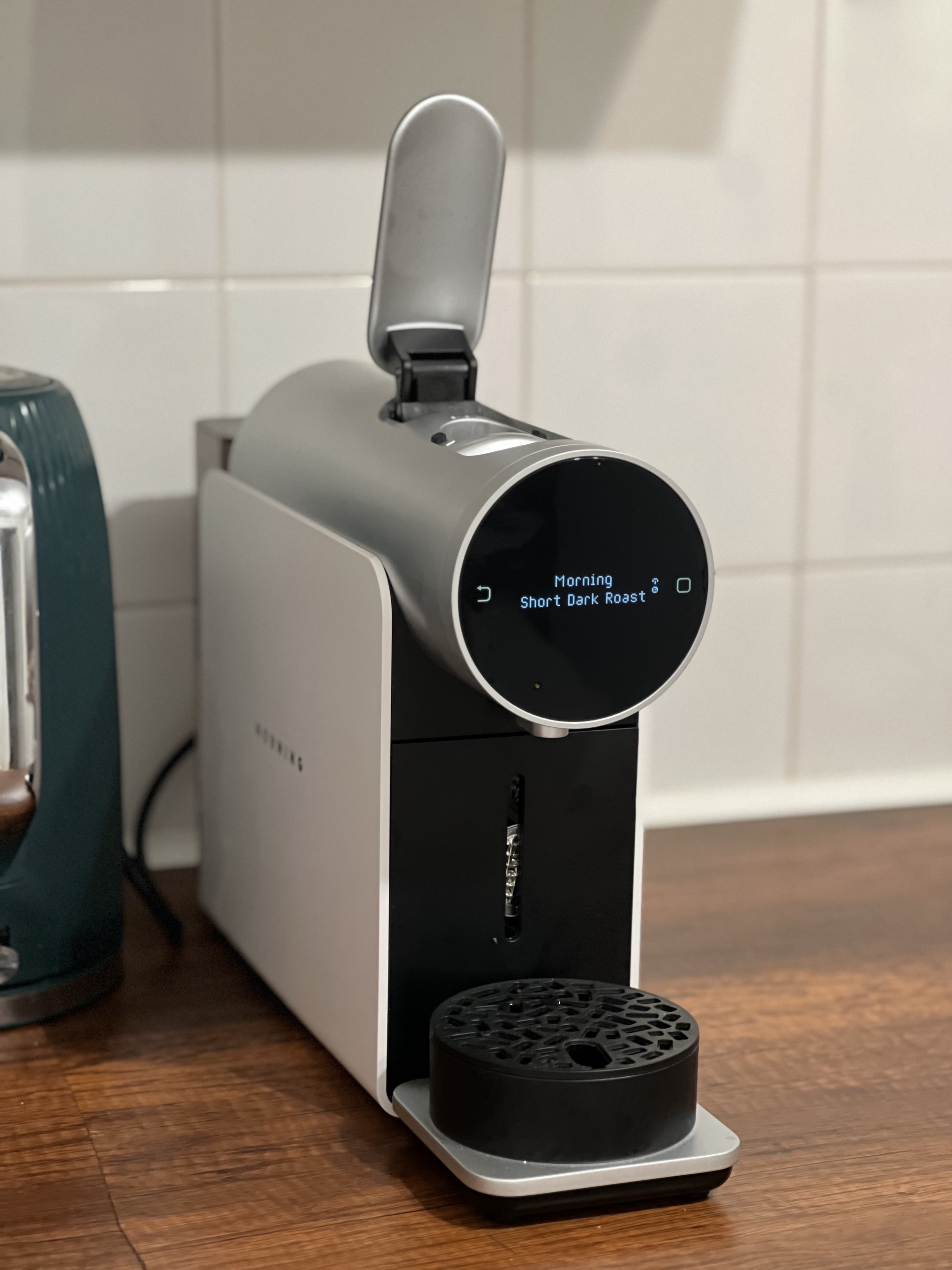 The Morning coffee pod machine in white on a wooden countertop that has a white backsplash.