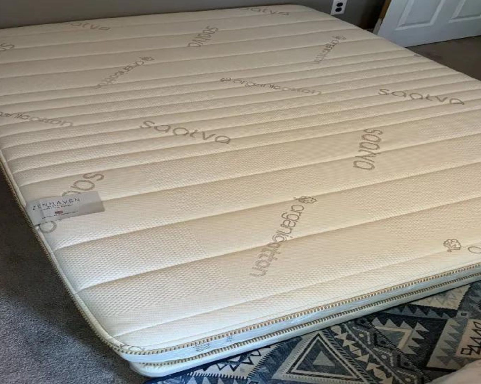 The Saatva Zenhaven mattress shown on the floor with a patterned blue rug beneath.