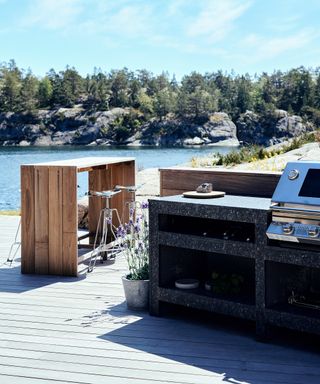 Garden bar ideas with a barbecue and wooden bar on pale gray decking in front of a lake with rocks and trees.