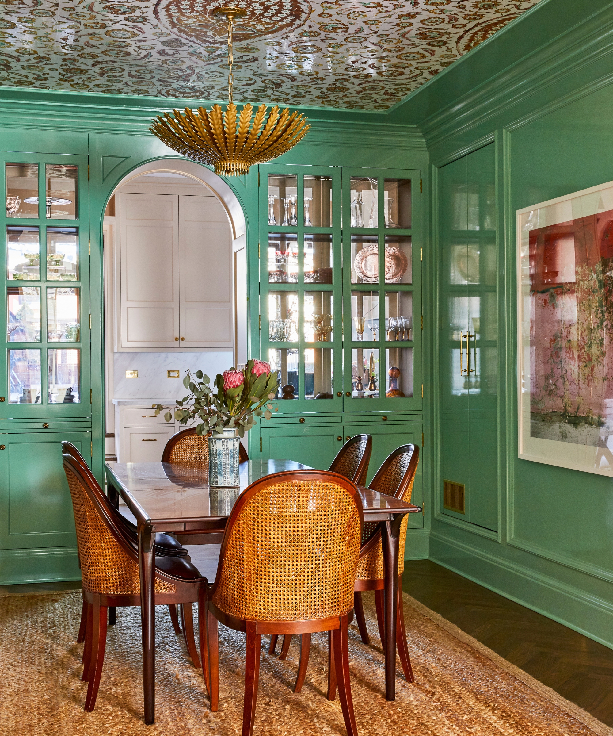 An emerald green dining room connected to a pale pink kitchen with double side glazed cabinets holding China and glassware