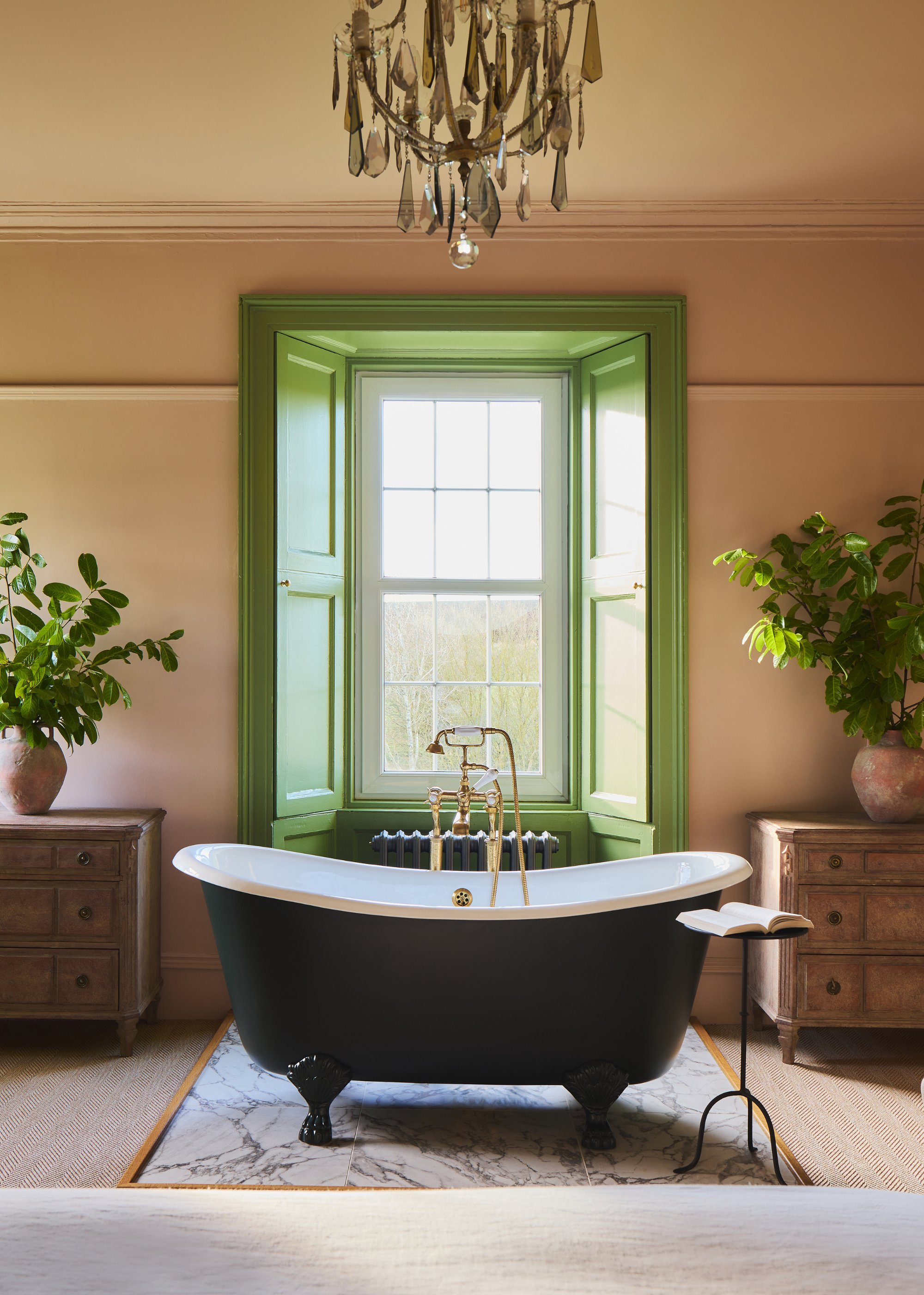 A primary bedroom with a freestanding bathtub with a green painted window recess that frames it with chests of drawers on either side of the window, with large vases filled with foliage and a chandelier hanging above the bath
