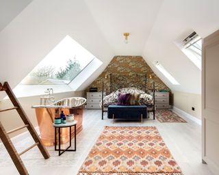 Loft style bedroom with vaulted ceiling, two skylights, wallpapered wall behind bed, metal bedframe, two patterned rugs, copper bath beside skylight,