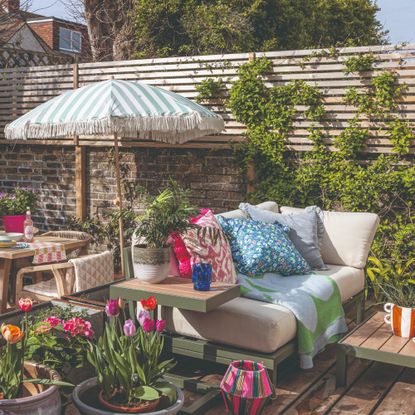 A garden with an outdoor sofa adorned with colourful outdoor cushions set next to a dining area with a striped fringed parasol