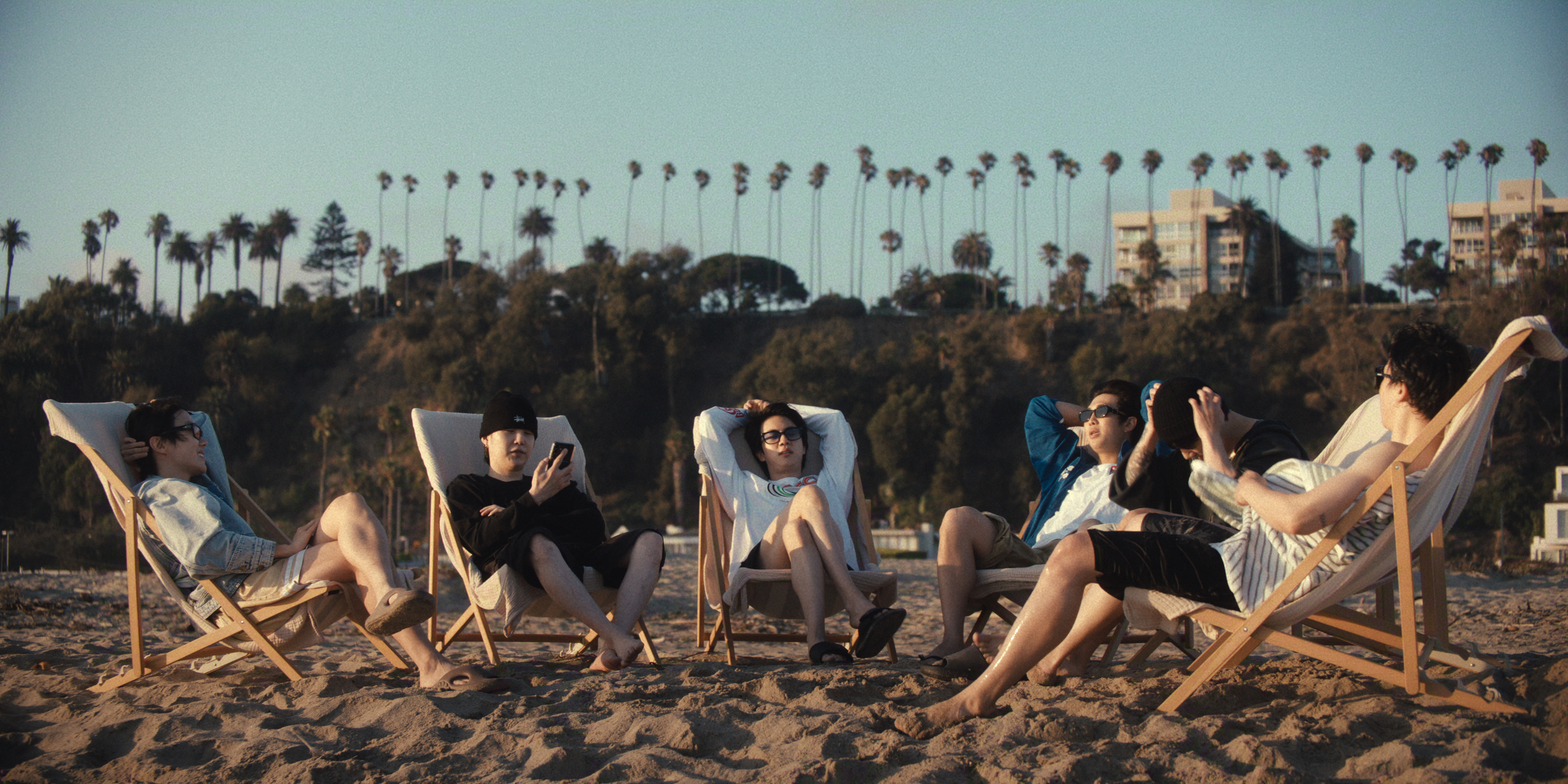 Members of BTS (l-r: j-hope, Suga, Jin, RM, Jung Kook, and Jimin) lounge in chairs on a Los Angeles beach, in the Netflix documentary 'BTS: THE RETURN.'