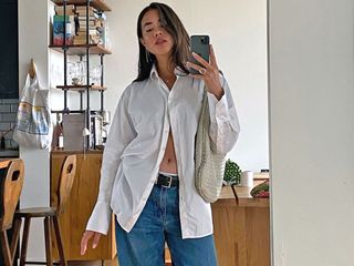 Fashion influencer Sasha Mei poses for a mirror selfie wearing a white button-down shirt, cream Bottega Veneta shoulder bag, black belt, and jeans