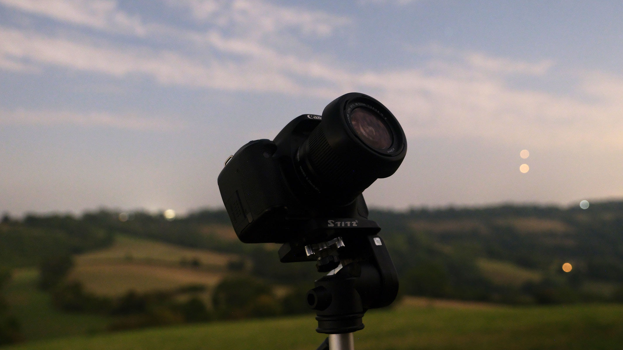 A Canon EOS Rebel T3i with a Canon EF-S 18-55mm f/3.5-5.6 IS II lens attached pointing at the sky with fields in the background with stars Castor and Pollux in the sky.