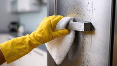 Hand in a yellow rubber glove using a white microfiber cloth to wipe moisture and water spots off a stainless steel appliance door and handle