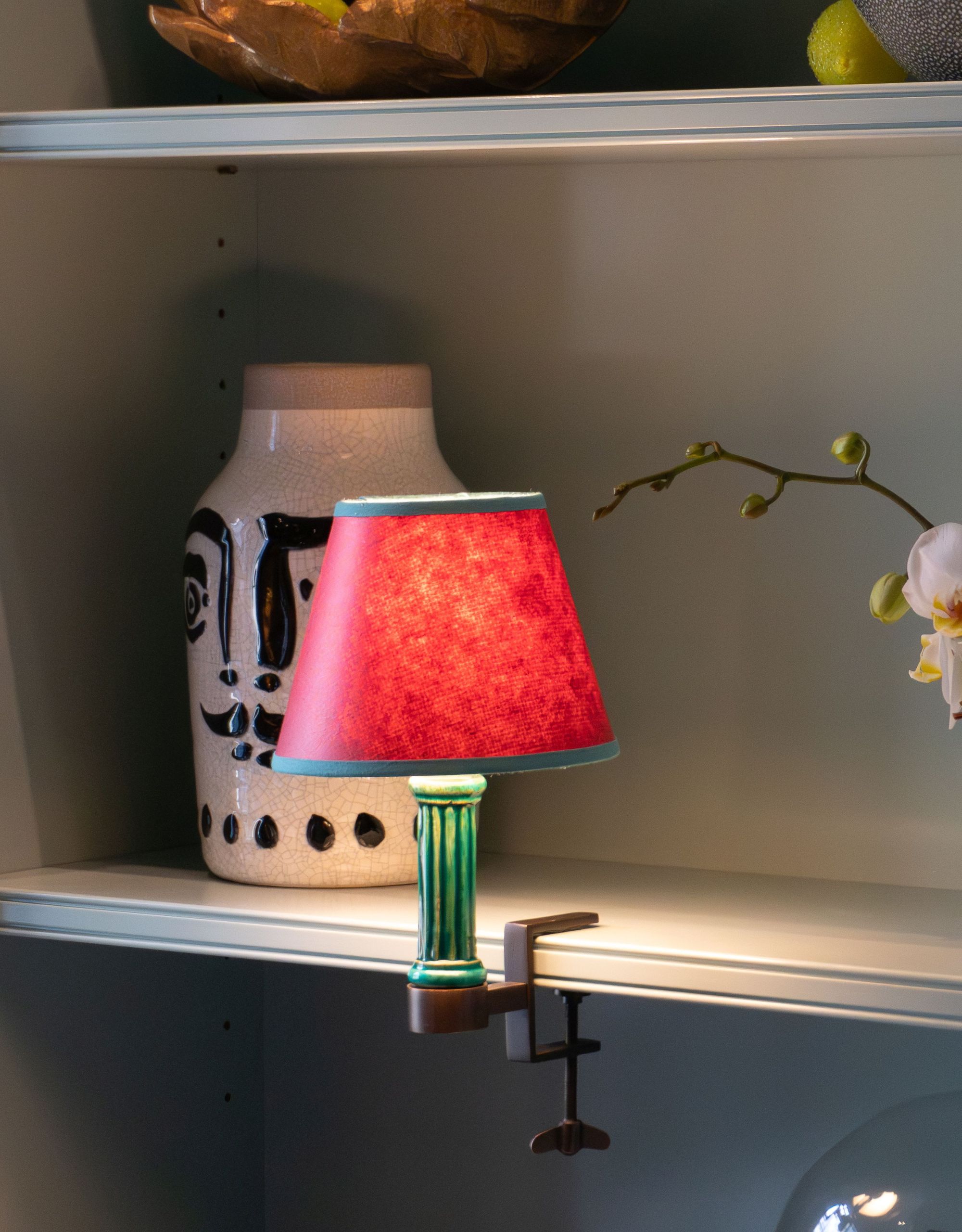 A bookshelf lamp with a green base and red shade with a green trim on a light-colored bookshelf with decorative objects