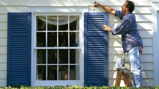 How to add value to your home: A man paints blue wooden shutters on a white house
