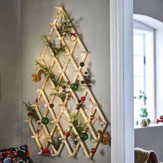 A gray wall with a wooden Christmas tree alternative hanging. There are green and red decorations and lights on the structure