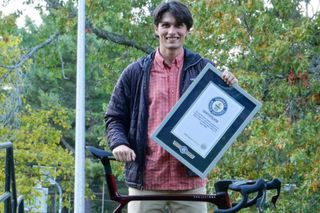 Sam Westby está detrás de su Trek Madone lateral en ropa de calle sosteniendo una ratificación enmarcada de su récord mundial Guinness.