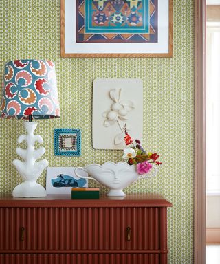Chest of drawers against a green and white wallpapered wall, lamp with coloured lampshade, framed picture and plinth above vase with face pattern.