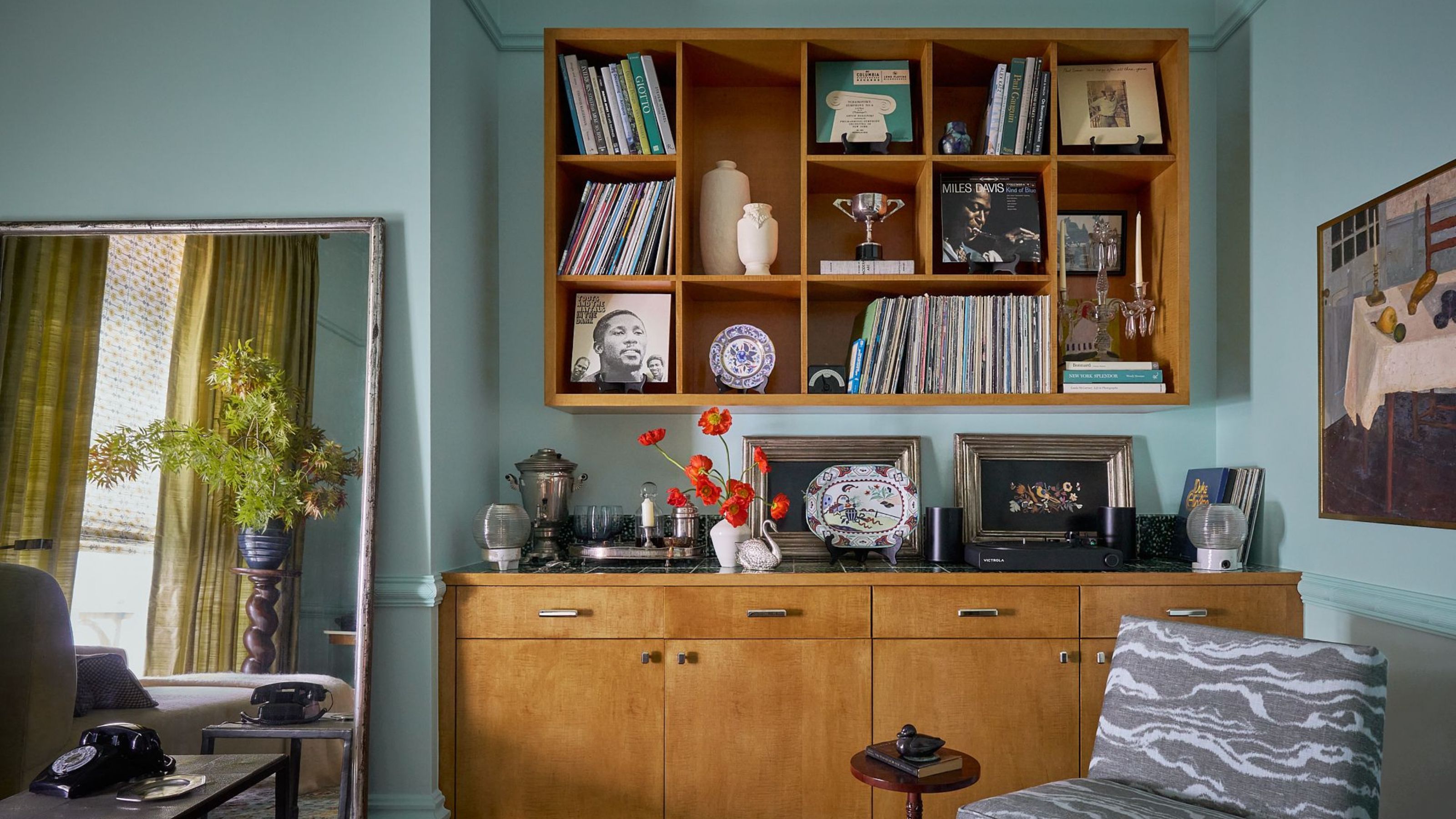 A living room with blue color-drenched walls, a large wooden cabinet with vintage decor, and a matching wooden wall unit above, with styled decor and books. 