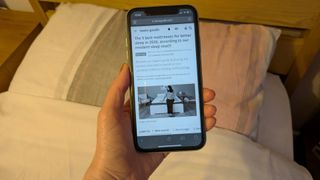 This image shows a hand holding an iPhone showing the Tom's Guide best mattress page in grayscale. In the background is a bed and pillows