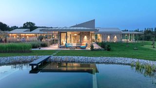 Contemporary single storey self build with metal roof and extensive glazing, surrounded by a landscaped garden with a large natural pool in front, shown at dusk