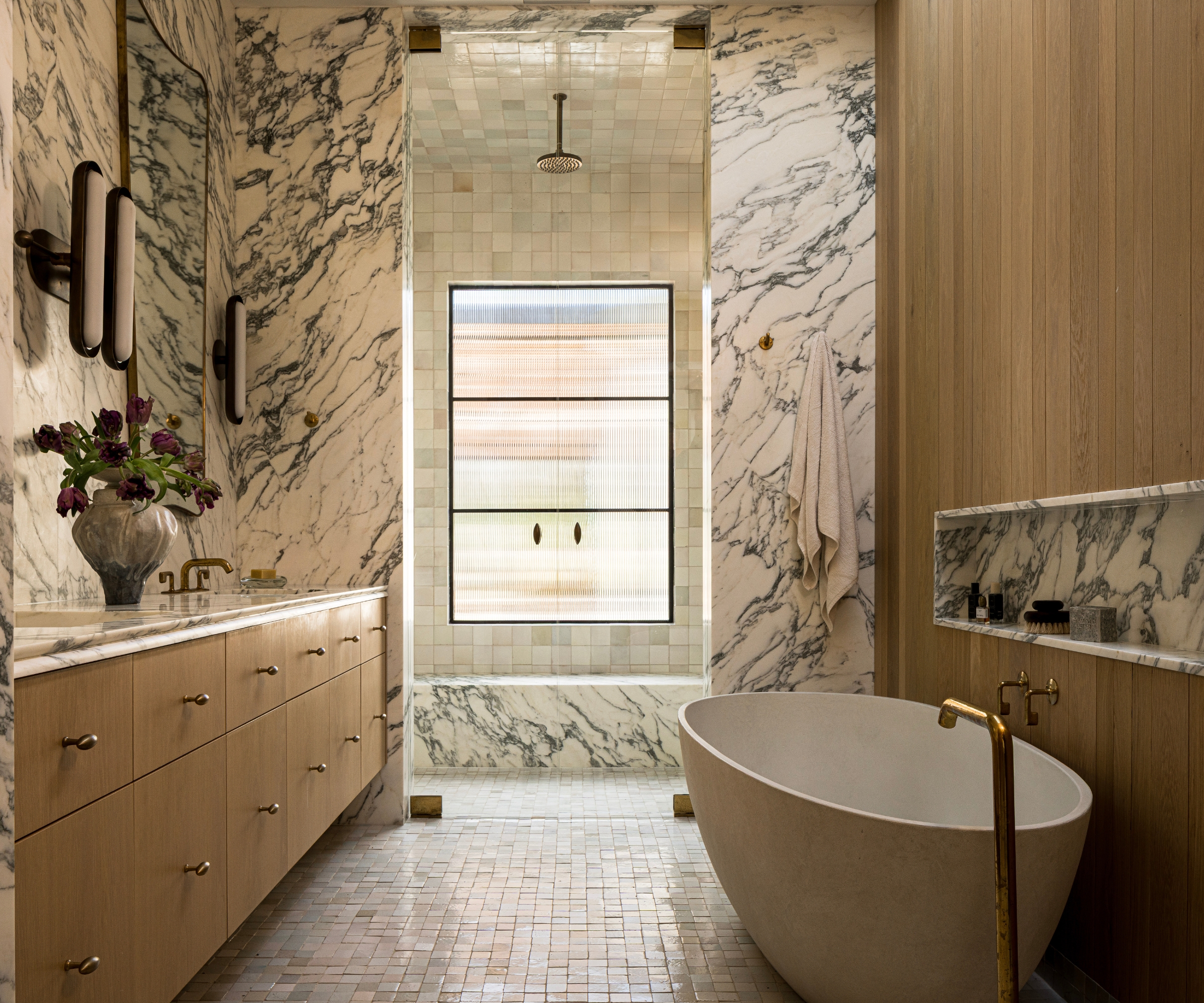 A large bathroom with warm wood finishes on the vanity and bath tub wall, and marble drenched across the walls and shower bench