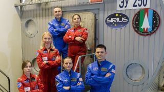 six crewmembers in spacesuits pose outside of a simulated spacecraft in a laboratory