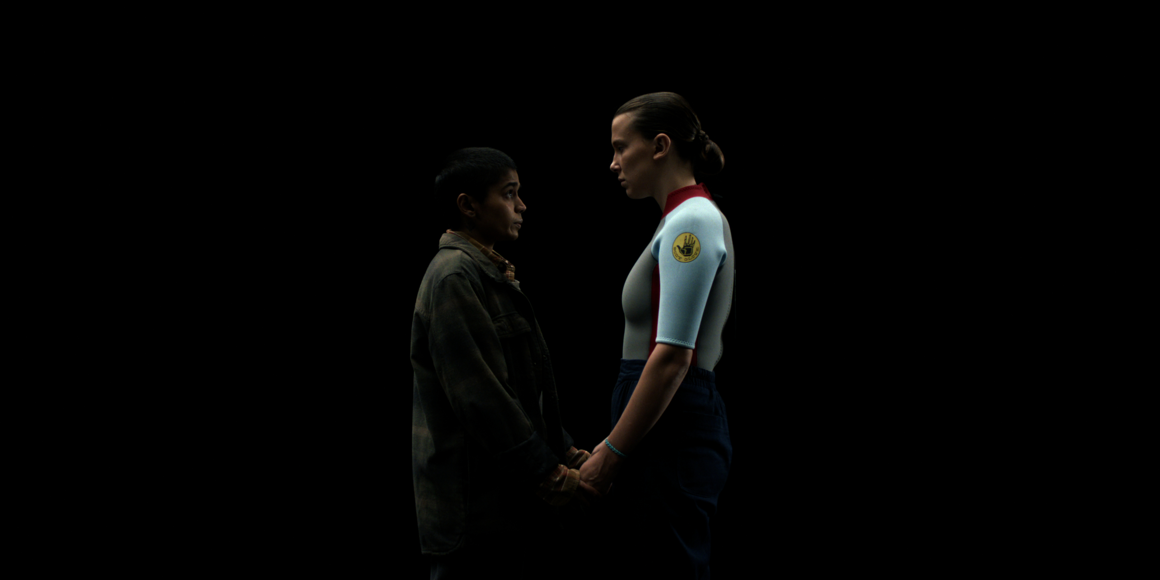 Linnea Berthelsen as Kali wearing a green military jacket and holding hands with Millie Bobby Brown as Eleven in a deprivation suit, standing in all black in the upside down in Stranger Things: Season 5