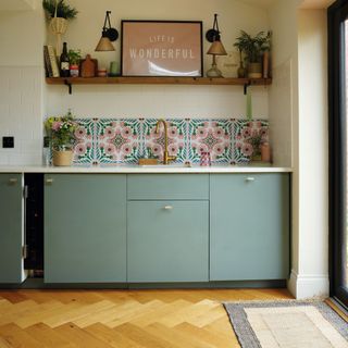 wooden parquered flooring, grey-green kitchen cabinets under patterned splashback and shelving