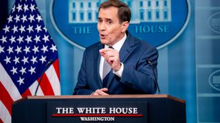 a man with short hair in a blue suit speaks into microphones at a lectern featuring a sign reading "the white house" in front of an american flag