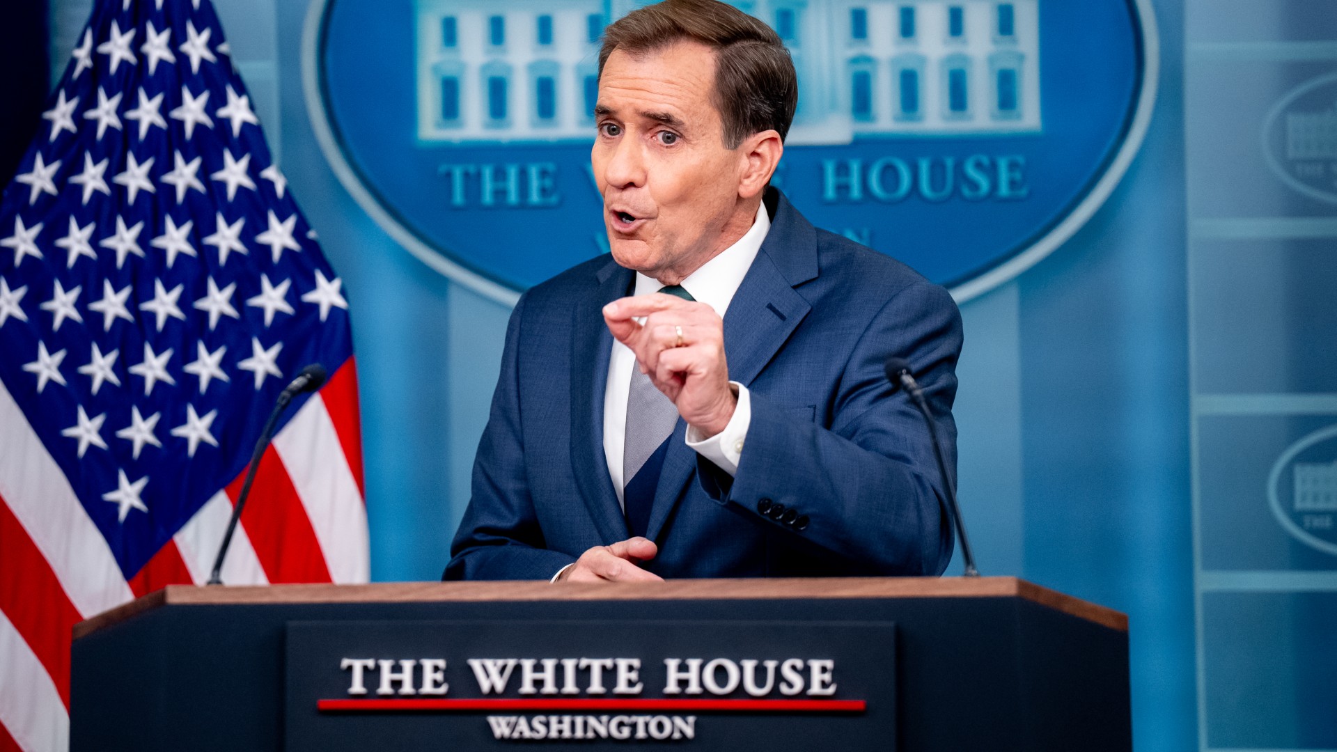 a man with short hair in a blue suit speaks into microphones at a lectern featuring a sign reading 