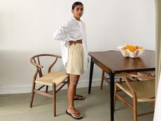 British style influencer Monikh Dale poses in her chic London dining room with an abstract white bowl full of oranges on the table wearing a white oversize line button-down shirt, butter-yellow shorts, anklets and black flat flip-flop sandals
