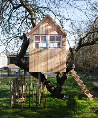 A wooden outdoor play hut with a ladder on top of a lawn, with a tree and a house behind it