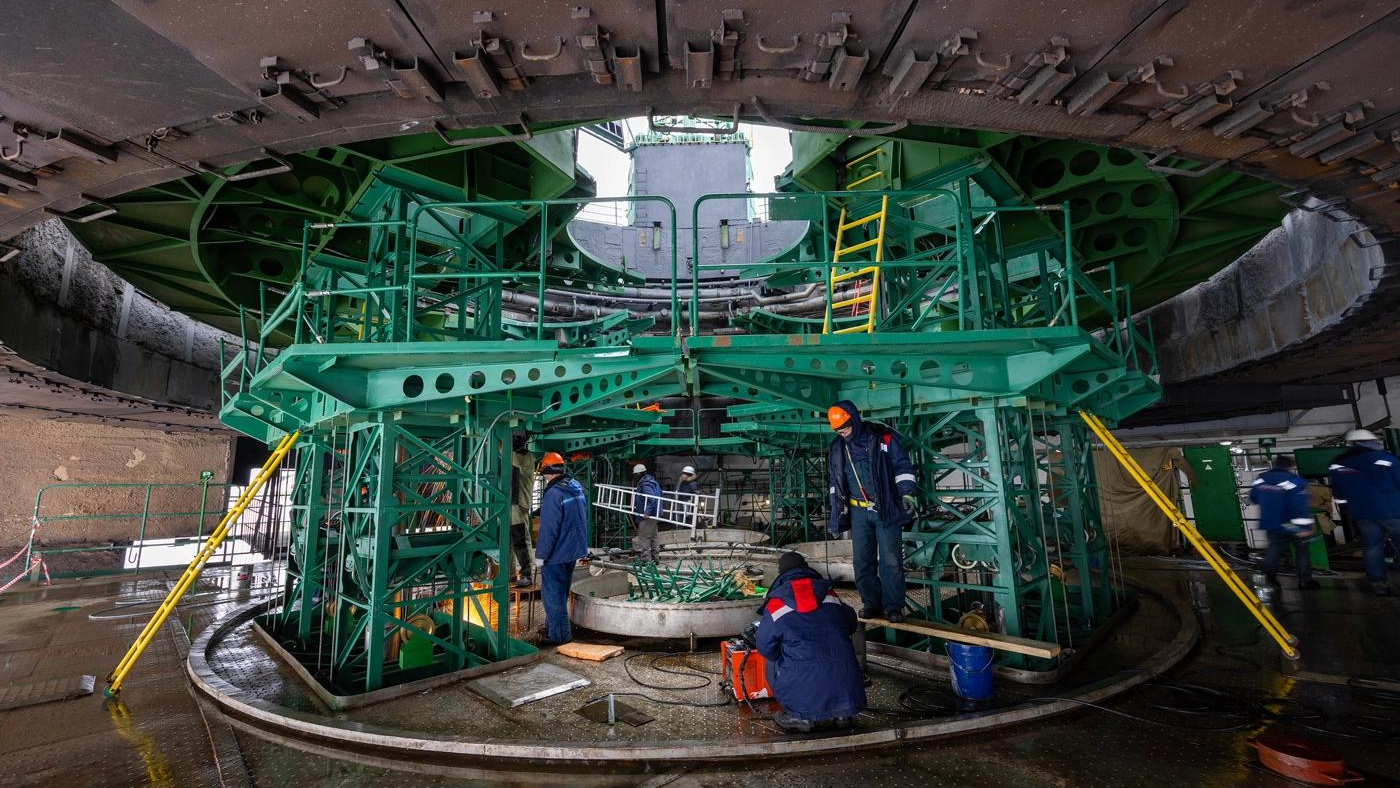 three workers in blue coveralls stand beneath a metallic green gantry