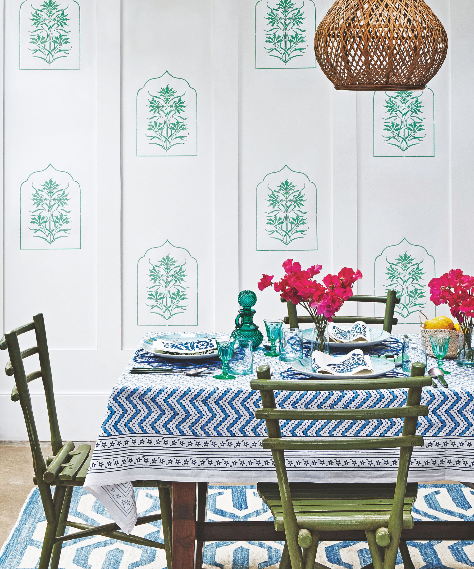 Turquoise and white dining room with blue and white patterned tablecloth, olive green wooden chairs, and vases of fuschias