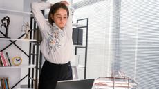 Woman stands in front of desk with open laptop stretching her arms behind her head