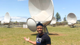 ramiro saide stands in front of a set of radio telescopes in a field with blue sky in behind