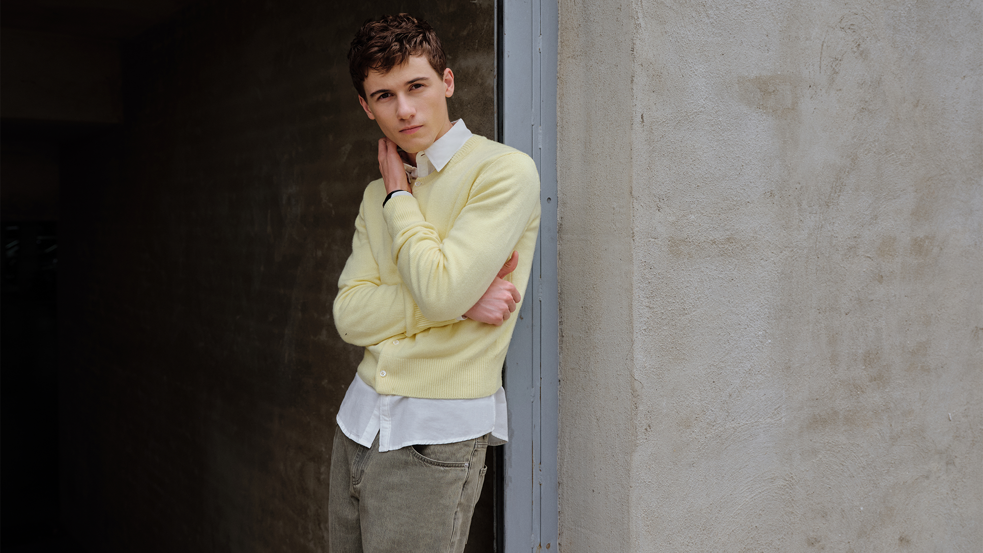 Sam Nivola, photographed by JJ Geiger. He is leaning against a cement wall with his arms crossed, wearing a butter-yellow cardigan over a white button-down shirt and gray jeans. 