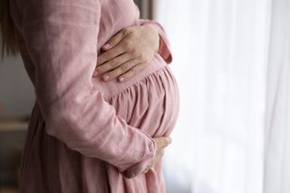 A pregnant woman in a pink dress holds her belly while looking out a window