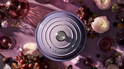 A purple lid to a Le Creuset Dutch oven viewed from above, on a purple worktop, surrounded by purple and light pink flowers