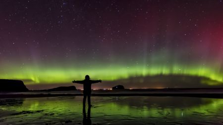 the silhouette of a woman standing on a beach with her arms outstretched, with a green aurora visible in the night sky