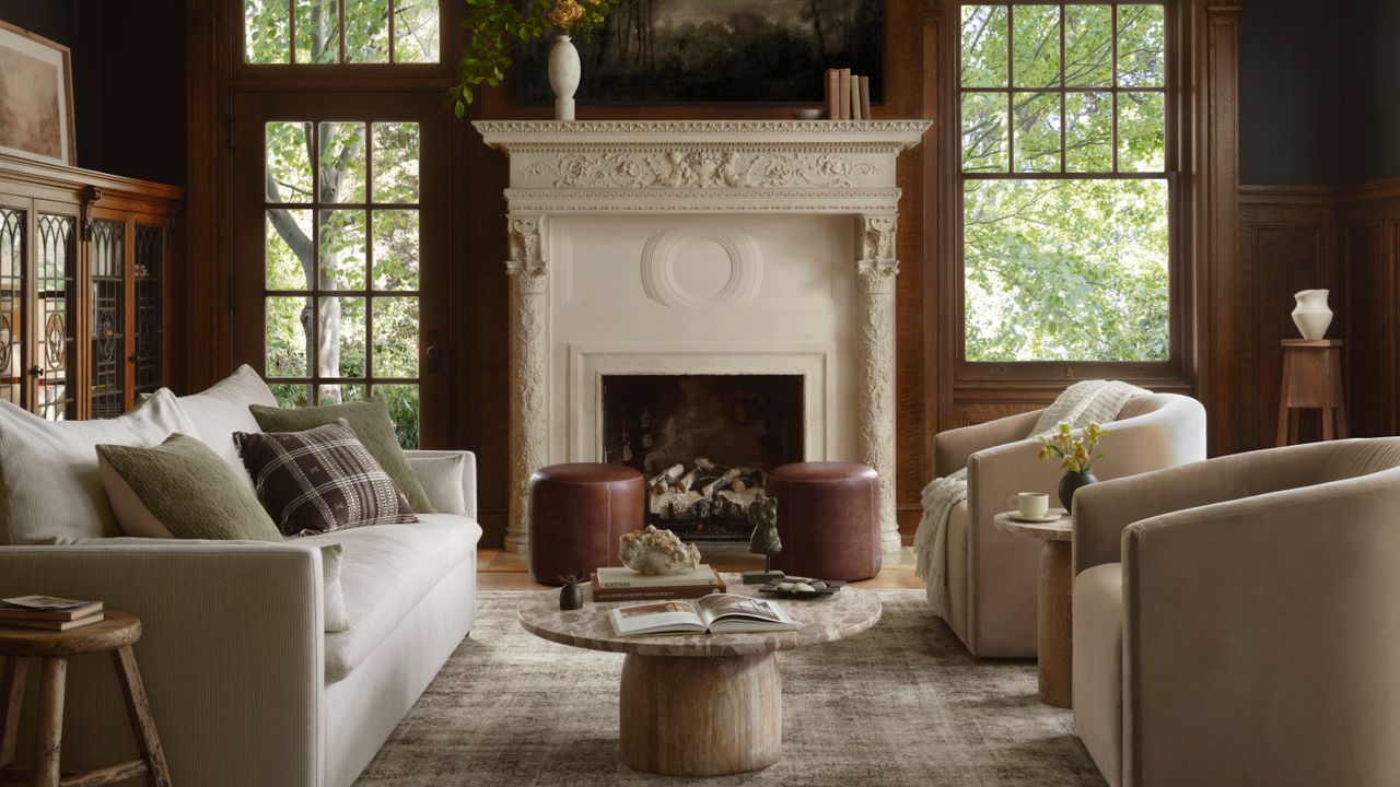 A living room with wooden walls and a grand off-white fireplace with windows on either side. There is a white sofa and two beige accent chairs facing each other and a grayish-green rug inbetween