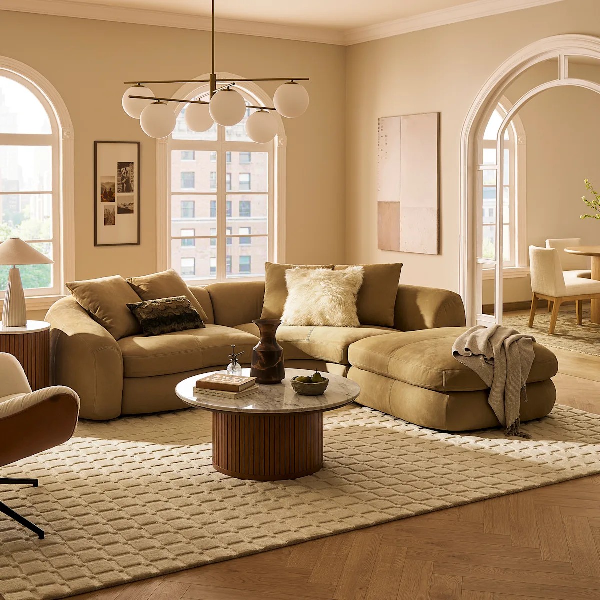 Image of a modern living room with a beige color scheme. There is an olive curved sofa with a small marble and wood coffee table in front of it. A tufted rug covers the floor.