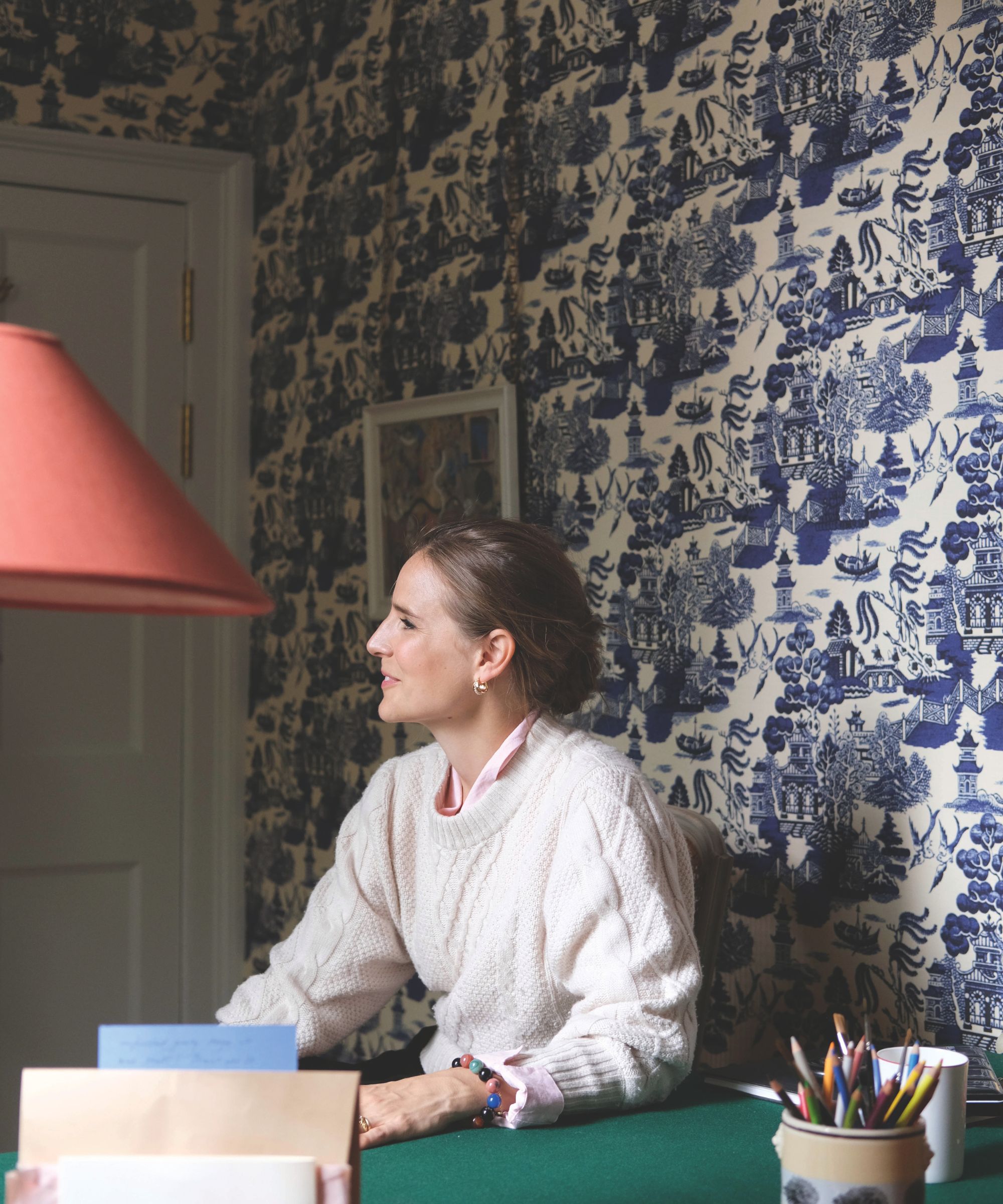 A side profile portrait of a woman in a pale pink jumper sits at a study desk. The wallpaper on the wall behind is a blue and white blue willow pattern.