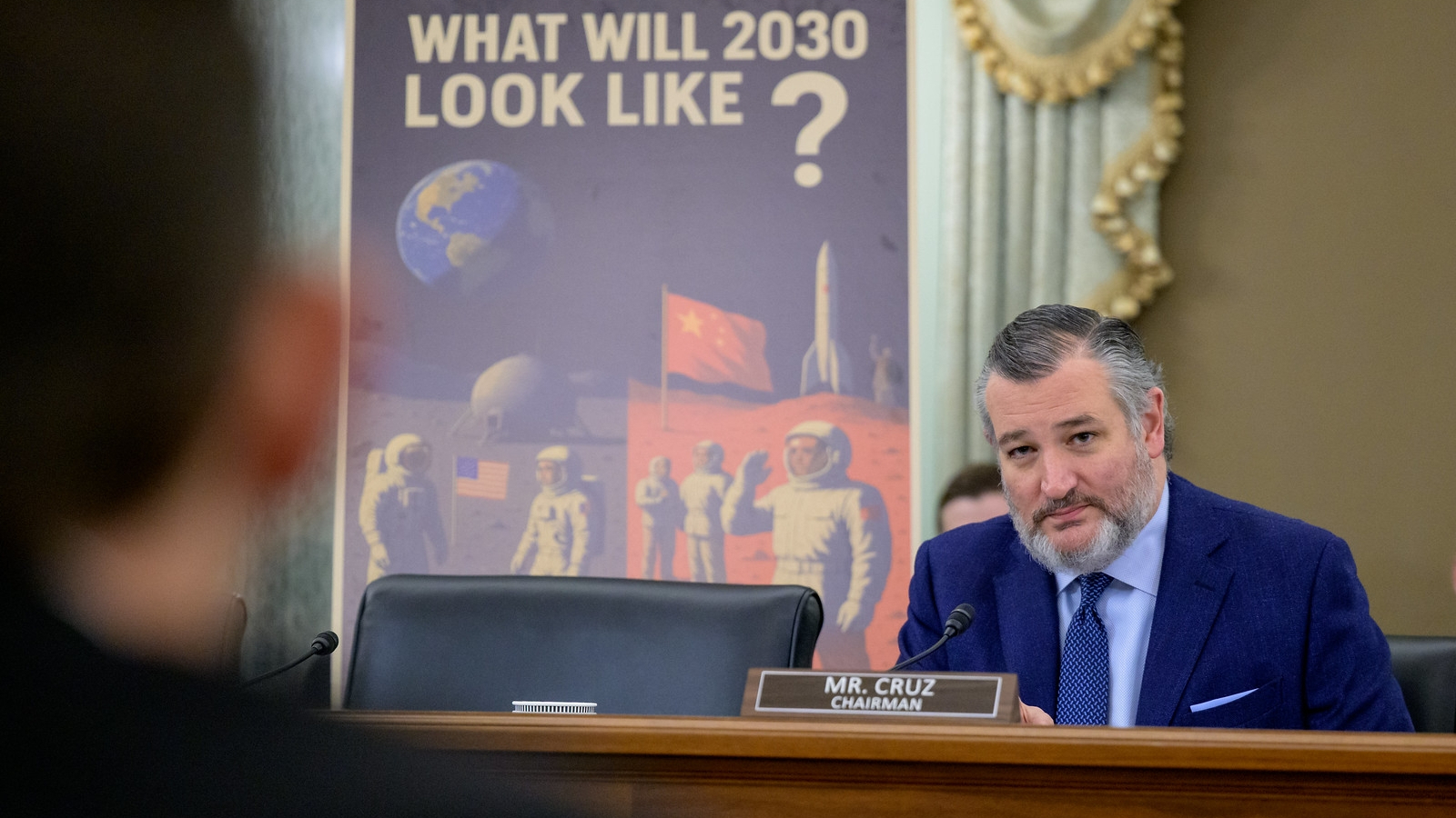 a bearded middle-aged man in a suit sits at a table