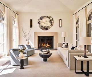living room with high ceiling and round mirror artwork over lit fire, two blue tub chairs and cream sofa