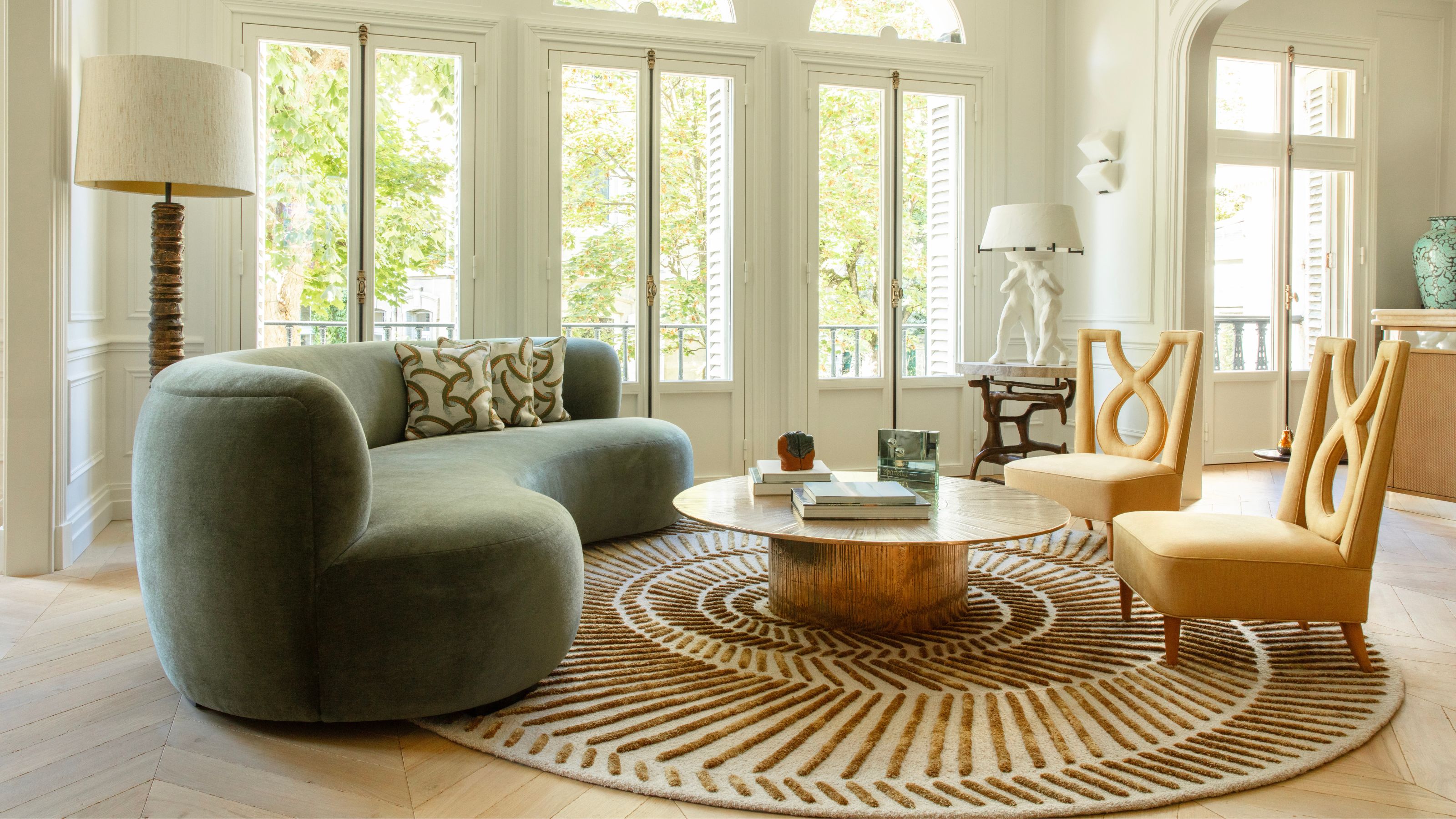 A bright living room featuring a curved sage green velvet sofa, a sunburst-patterned rug, and two sculptural yellow armchairs arranged around a low brass coffee table