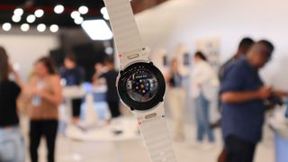 Samsung Galaxy Watch 7 held aloft with a busy room in the background. The rear is showing