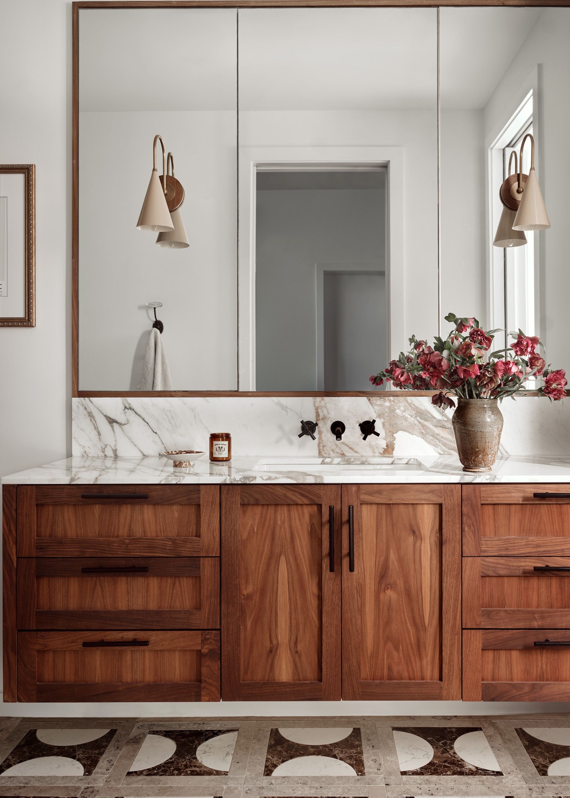 A bathroom with a large vanity with drawers and cabinets as well as a large mirror above it. On the bathroom countertop is a vase with pink flowers inside