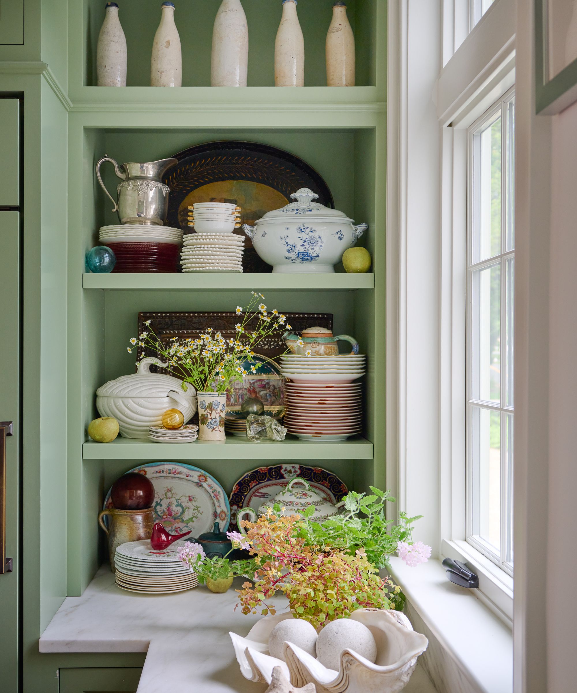 Kitchen with pale green shelves decorated with vintage china tea sets, plates, and bowls, and a marble countertop with fresh herbs and a shell bowl