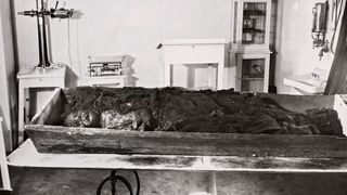 This black and white photo shows the remains of a young woman "bog body" sitting on a long wooden box on a table.